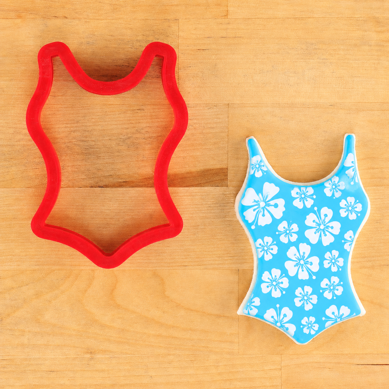 Red cookie cutter shaped like a swimsuit next to a blue swimsuit cookie, stenciled white floral cookie stencil, lying on a wooden surface
