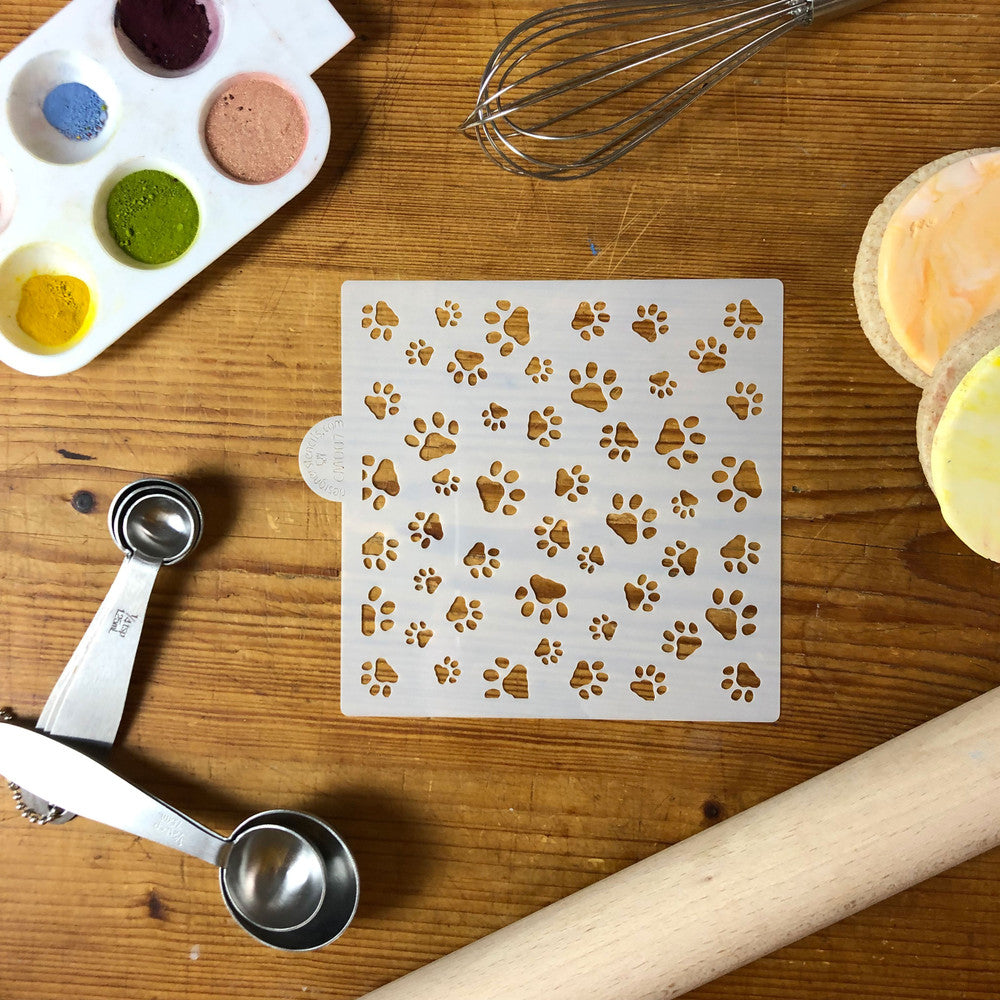 Mini dog paws cookie stencil on wooden table with measuring spoons, whisk, and colored powders.