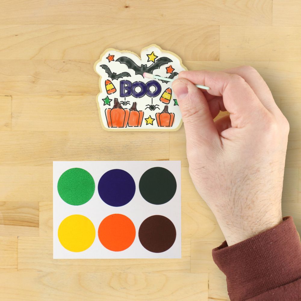 Hand painting a Halloween cookie with a 'BOO' stencil, bats, pumpkins, and colorful paint palette.