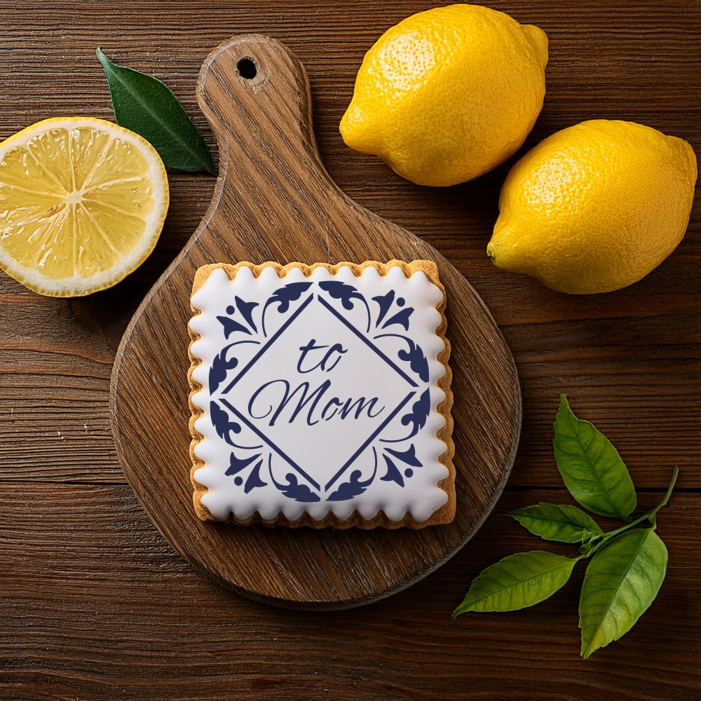 Mother's Day cookie decorated with 'To Mom' blue tile stencil, surrounded by lemons and leaves on a wooden board.