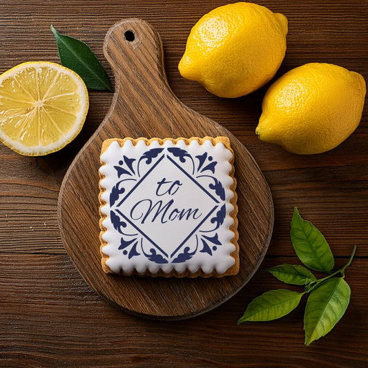 Mother's Day cookie decorated with 'To Mom' blue tile stencil, surrounded by lemons and leaves on a wooden board.