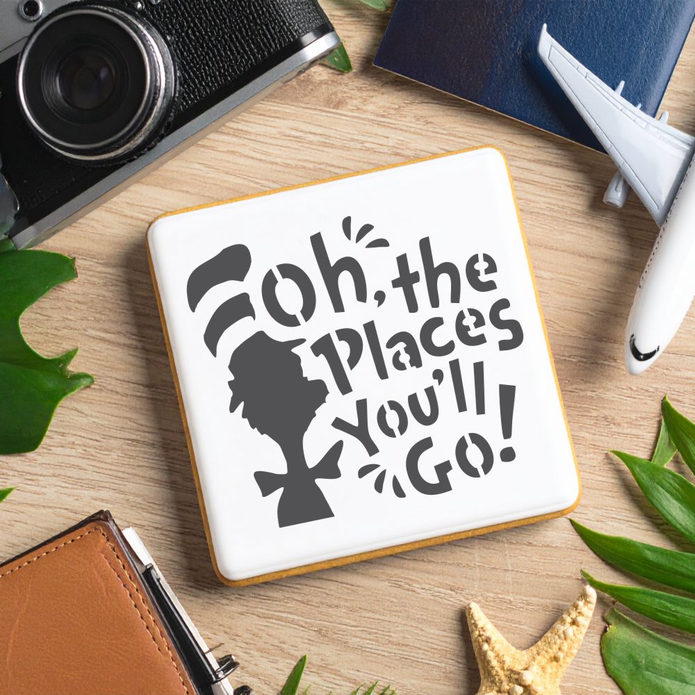 Cookie with 'Oh, the Places You'll Go!' stencil design on a wooden table surrounded by leaves and travel items.