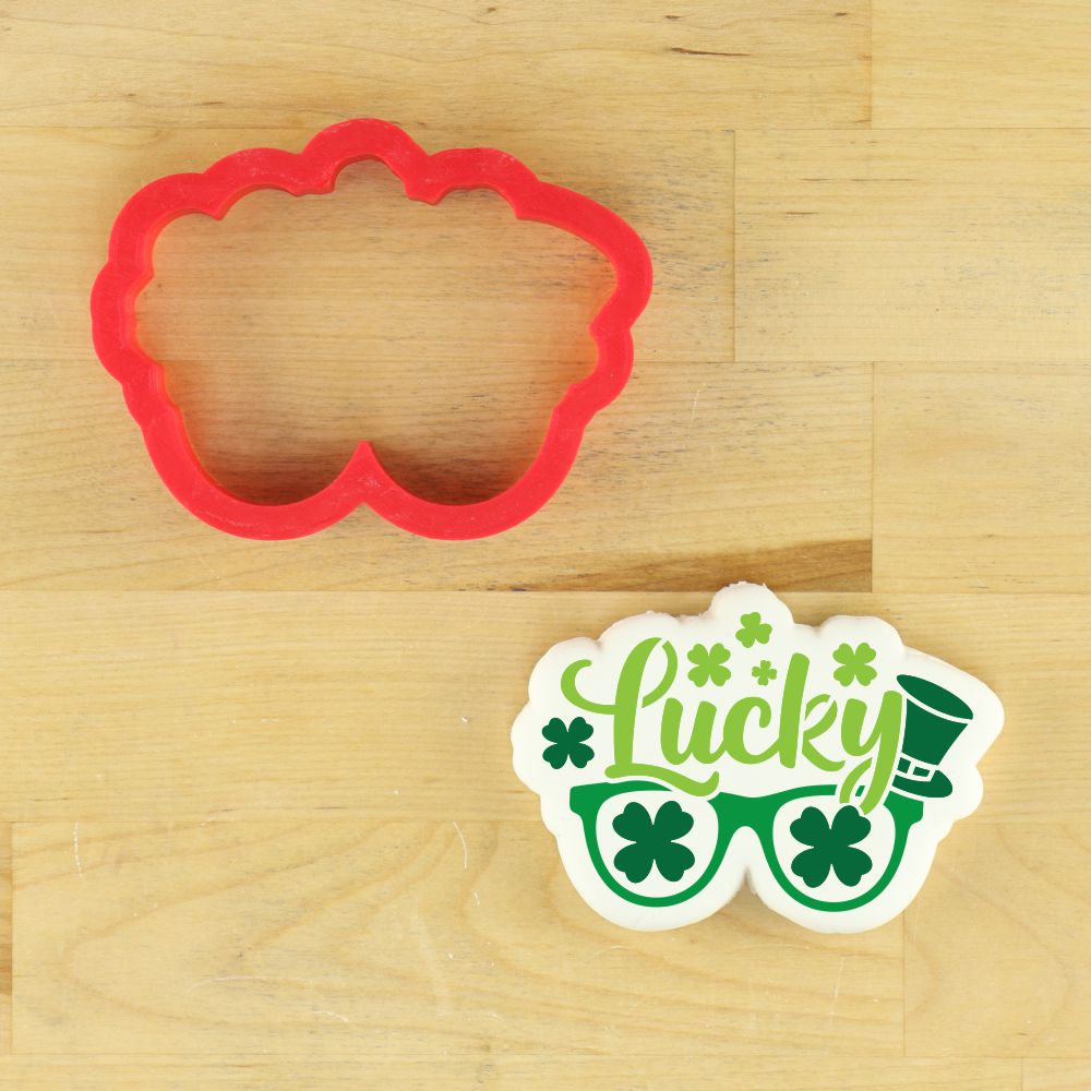 St. Patrick's Day sunglasses cookie stencil and matching red cookie cutter on a wooden surface.