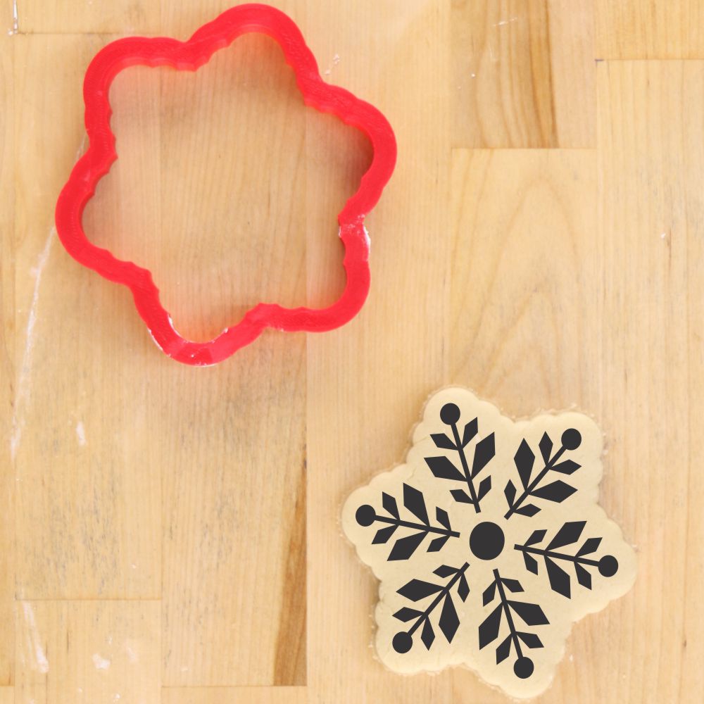 Red snowflake cookie cutter beside a decorated snowflake cookie stencil on a wooden surface.