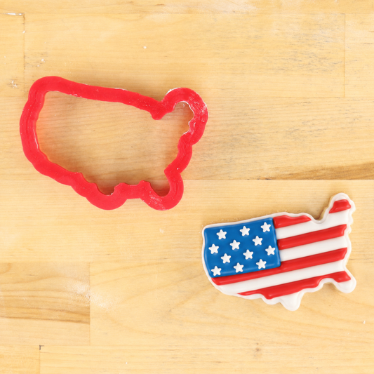 A red plastic cookie cutter shaped like the outline of the United States is shown next to a small decorated sugar cookie in the shape of the USA with an American flag design, placed on a light wood surface.