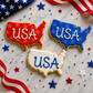 Three cookies shaped like the United States with 'USA' text, surrounded by American flags and stars on a marble surface.