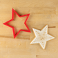 A red five-point star-shaped cookie cutter and a decorated star-shaped cookie with gold sprinkles on a light wood background.