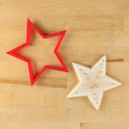 A red five-point star-shaped cookie cutter and a decorated star-shaped cookie with gold sprinkles on a light wood background.