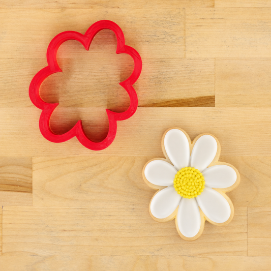 A red flower-shaped cookie cutter placed on a light wood surface next to a decorated daisy-shaped cookie with white petals and a yellow center.