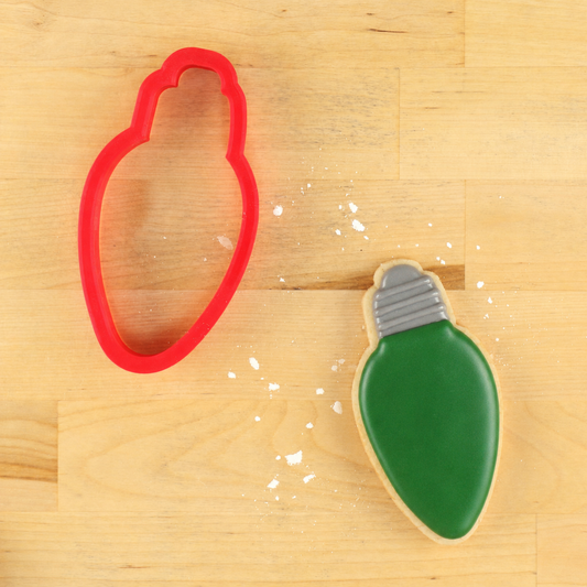 A red holiday light-shaped cookie cutter and a decorated light-bulb-shaped sugar cookie on a light wood surface.