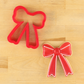 Christmas bow cookie cutter next to a ribbon-shaped cookie decorated with red flooded icing for the holidays