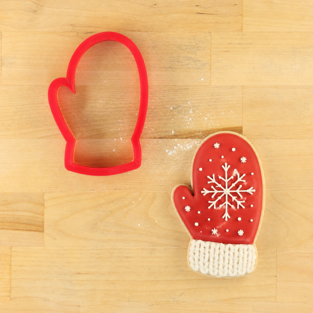A red mitten-shaped cookie cutter shown next to a decorated red mitten cookie with white icing snowflakes and a white knitted cuff, placed on a light wood surface.