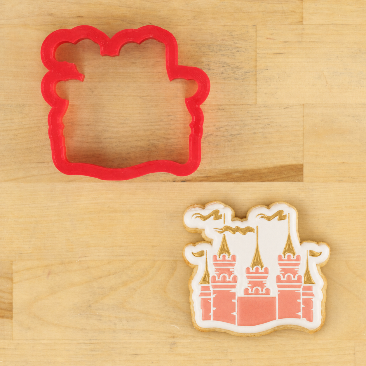 A red cookie cutter shaped like a castle and a decorated sugar cookie featuring a castle design, displayed on a light wood background.