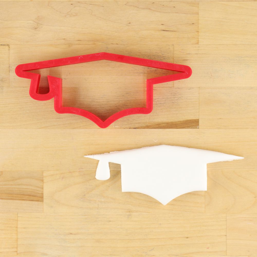 Red graduation cap cookie cutter on a wooden surface with a black and white graduation cap decoration.
