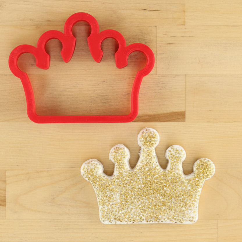 Crown shaped cookie cutter with a crown shaped cookie next to it iced, and topped with gold glitter.