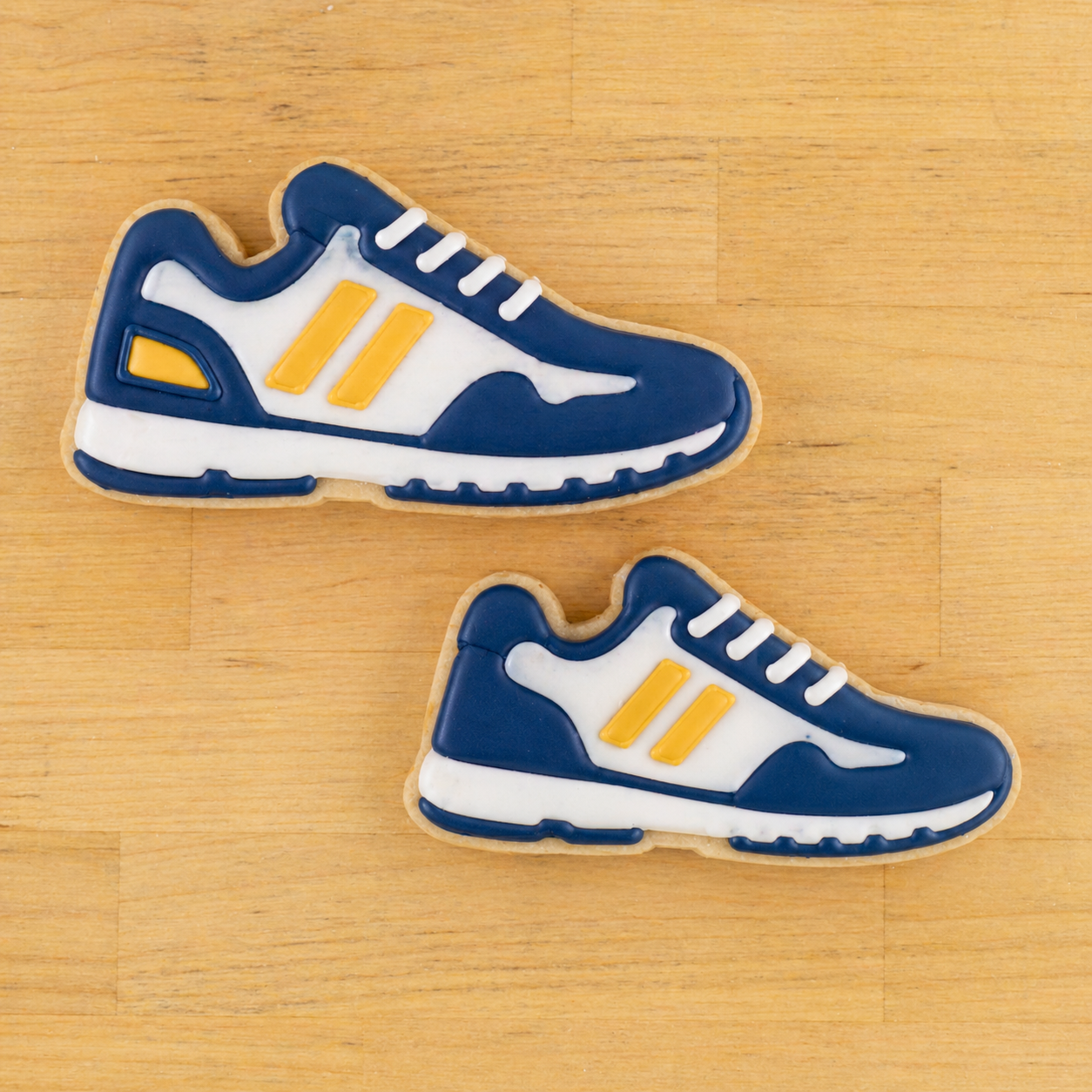 Two cookies shaped like blue and white sneakers with yellow accents on a wooden surface.