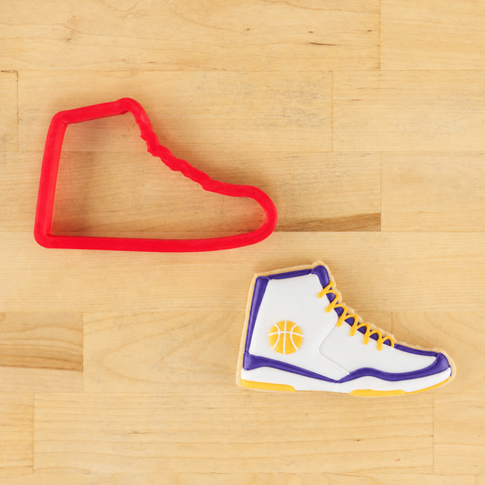 Cookie cutter shaped like a basketball shoe next to a cookie decorated like a basketball shoe on a wooden surface