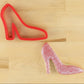 Red high-heeled shoe cookie cutter next to a pink glittery cookie shaped like a high-heeled shoe on a wooden surface.