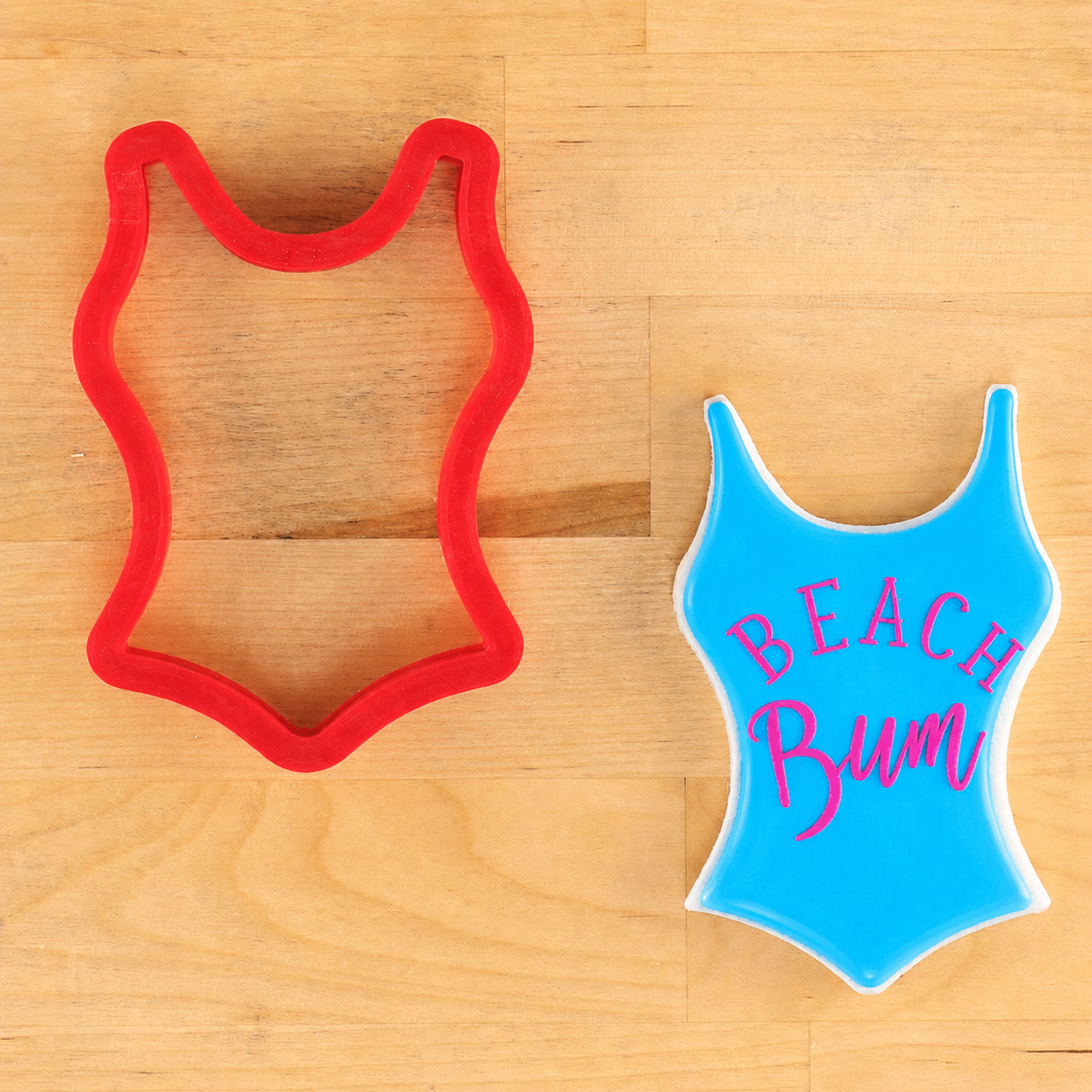 Red cookie cutter shaped like a swimsuit and blue swimsuit with 'BEACH Bum' text on a wooden surface.