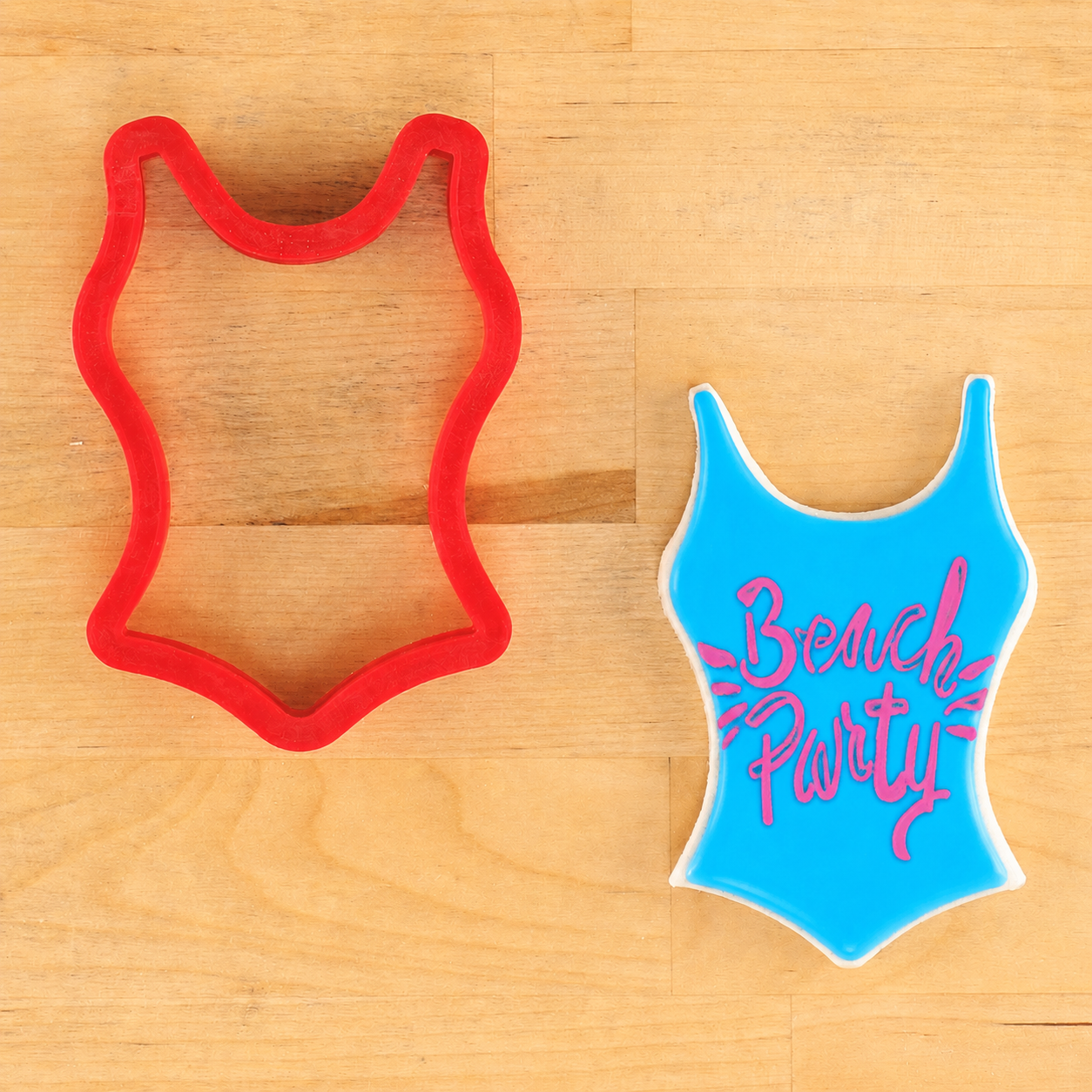Red cookie cutter shaped like a swimsuit and blue swimsuit with 'Beach Party' text on a wooden surface.