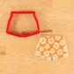 Red cookie cutter shaped like swim trunks next to an orange iced cookie with white floral patterns stencil onto it.