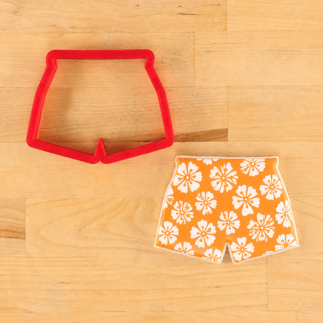 Red cookie cutter shaped like swim trunks next to an orange iced cookie with white floral patterns stencil onto it.