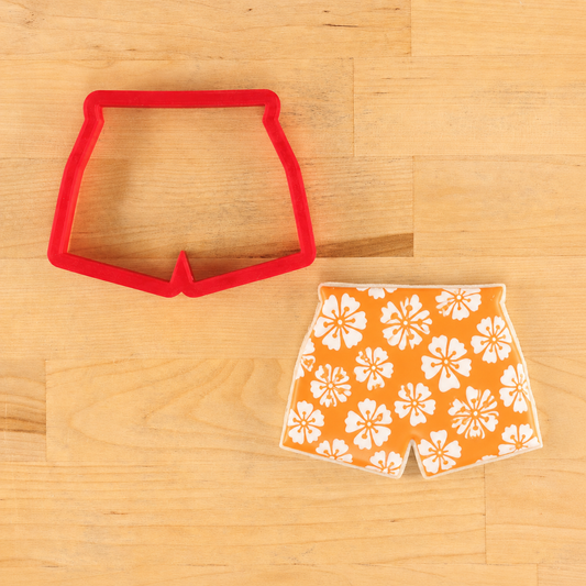 Red cookie cutter shaped like swim trunks next to an orange iced cookie with white floral patterns stencil onto it.