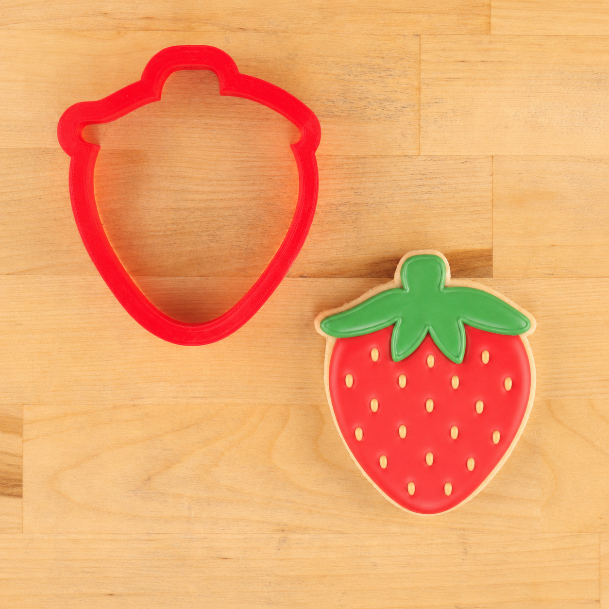 Red strawberry-shaped cookie cutter next to a strawberry-shaped cookie on a wooden surface