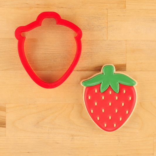 Red strawberry-shaped cookie cutter next to a strawberry-shaped cookie on a wooden surface