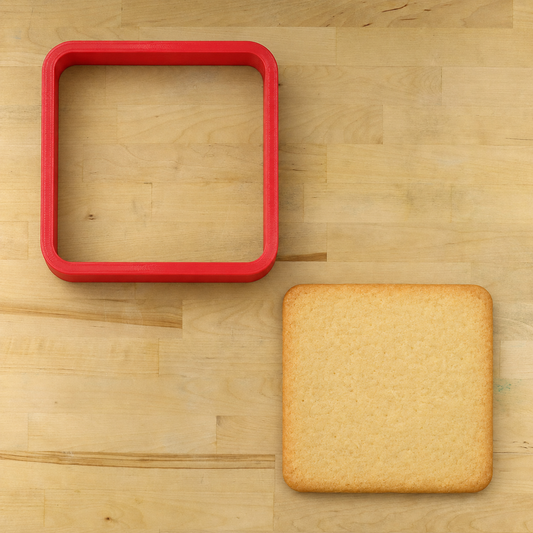 A square red silicone cookie cutter shown on a light wood surface next to a baked square cookie or biscuit used as a size reference.