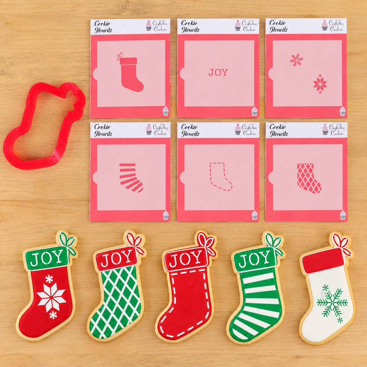 A set of Christmas-themed cookie cutters and decorated sugar cookies arranged on a light wood background. The set includes a red stocking-shaped cutter, six cookie stencils with multi-design overlays, and five finished cookies decorated in red, green, and white with 'JOY' text and snowflake patterns.