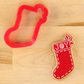 A red Christmas stocking-shaped cookie cutter and a decorated stocking-shaped cookie on a light wood background. The cookie is red with white dashed piping and a white cuff reading 'JOY' in white letters.