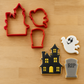 A set of Halloween-themed cookie cutters in red shown with three decorated sample cookies on a light wood background: a haunted house, a ghost, and a tombstone.