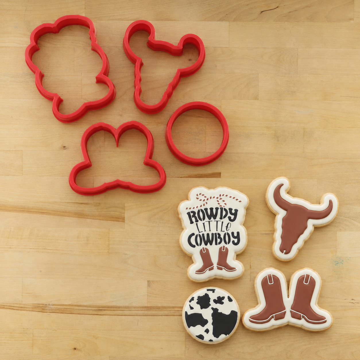 Cowboy Birthday party stencil and cutter set being used on iced and decorated sugar cookies featuring cowboy and cow motifs, including boots, a longhorn, a cow-print cookie, and a sign that reads 'Cowboy Little Cowboy'.