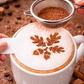 Cappuccino with cinnamon snowflake design being added by a hand using a small sieve.