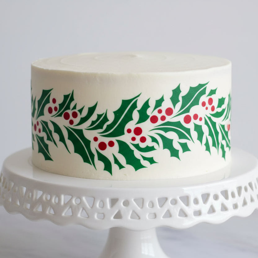White cake with holly leaf and berry design on a white stand against a light gray background