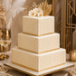 Three-tiered square wedding cake with gold and white pattern, floral decorations on a decorative table.