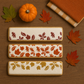 Three rectangular white cookies decorated with autumn leaf patterns in red, orange, and gold, arranged on a wooden surface with a small pumpkin, fall leaves, and a folded orange-and-beige striped cloth.