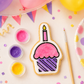 A decorated cupcake-shaped sugar cookie with pink and purple icing sits on a light surface with small paint pots, a paintbrush, balloons, confetti, and party decorations around it.