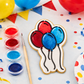 A decorated sugar cookie shaped like balloons, shown on a party-themed tabletop with small paint cups, a paintbrush, balloons, bunting, and confetti.