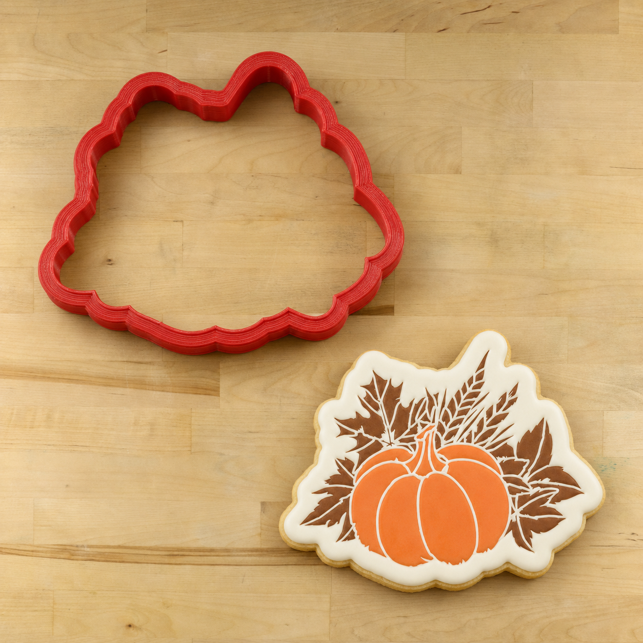 White iced cookie with stenciled pumpkin and leaves lying next to its matching cookie cutter