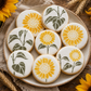 Nine round sugar cookies decorated with white icing and yellow sunflower motifs, arranged on a beige ceramic plate with wheat stalks and sunflower petals around it.