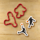 2 Red basketball player cookie cutters, lying next to two basketball player cookies, that have been flooded in white icing, then stenciled with the basketball player cookie stencils.