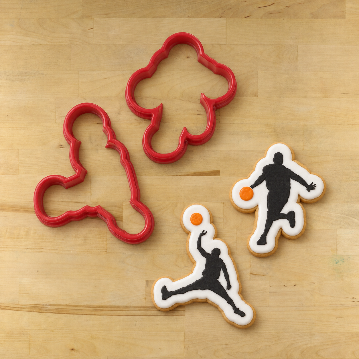 2 Red basketball player cookie cutters, lying next to two basketball player cookies, that have been flooded in white icing, then stenciled with the basketball player cookie stencils.