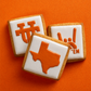 Three square iced cookies arranged on a bright orange background. Each cookie has white icing and an orange graphic: a Texas state outline, a stylized 'T' logo, and a hand gesture with 'HOOK 'EM'.