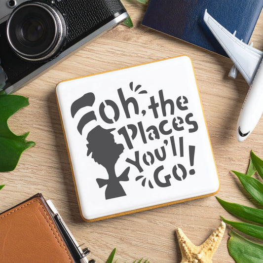 Cookie with 'Oh, the Places You'll Go!' stencil design on a wooden table surrounded by leaves and travel items.