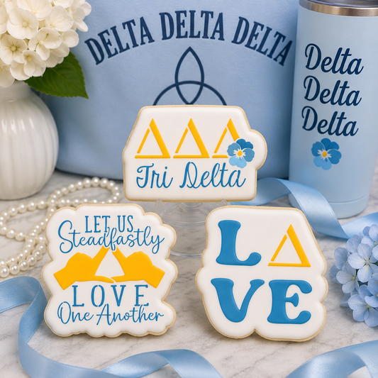 Three decorated Delta Delta Delta sugar cookies displayed with light blue sorority-themed décor, ribbons, pearls, and floral accents.