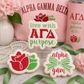 Three decorated Alpha Gamma Delta sugar cookies displayed with pink and green sorority-themed décor, ribbons, pearls, and floral accents.