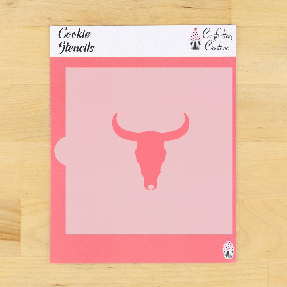 A pink cookie stencil sheet with a bull skull cutout design, displayed on a light wood background. The stencil packaging header reads 'Cookie Stencils' and features the 'Confederation of Confectioners' logo.