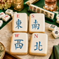 Four rectangular white cookies decorated like mahjong tiles with blue and red Chinese characters and letter markers, arranged on a stone board with real mahjong tiles and drink glasses in the background.